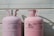 Two large, pink Dynatemp gas cylinders stand side by side in front of a white metal wall. The cylinders display branding and text information in black, including details such as 'Dynatemp 410A.' Their tops are slightly open with visible valves.