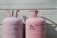 Two large, pink Dynatemp gas cylinders stand side by side in front of a white metal wall. The cylinders display branding and text information in black, including details such as 'Dynatemp 410A.' Their tops are slightly open with visible valves.