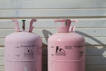 Two large, pink Dynatemp gas cylinders stand side by side in front of a white metal wall. The cylinders display branding and text information in black, including details such as 'Dynatemp 410A.' Their tops are slightly open with visible valves.