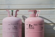 Two large, pink Dynatemp gas cylinders stand side by side in front of a white metal wall. The cylinders display branding and text information in black, including details such as 'Dynatemp 410A.' Their tops are slightly open with visible valves.