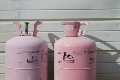 Two large, pink Dynatemp gas cylinders stand side by side in front of a white metal wall. The cylinders display branding and text information in black, including details such as 'Dynatemp 410A.' Their tops are slightly open with visible valves.