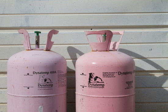 Two large, pink Dynatemp gas cylinders stand side by side in front of a white metal wall. The cylinders display branding and text information in black, including details such as 'Dynatemp 410A.' Their tops are slightly open with visible valves.