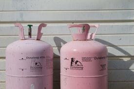 Two large, pink Dynatemp gas cylinders stand side by side in front of a white metal wall. The cylinders display branding and text information in black, including details such as 'Dynatemp 410A.' Their tops are slightly open with visible valves.