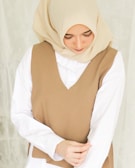 A woman wearing a beige hijab and a brown sleeveless vest over a white shirt. She is looking down with a serene expression, and her hands are gently clasped in front of her.