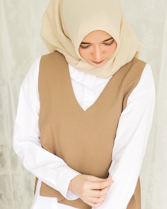 A woman wearing a beige hijab and a brown sleeveless vest over a white shirt. She is looking down with a serene expression, and her hands are gently clasped in front of her.