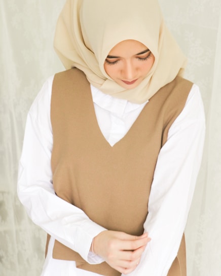 A woman wearing a beige hijab and a brown sleeveless vest over a white shirt. She is looking down with a serene expression, and her hands are gently clasped in front of her.