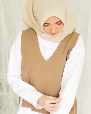 A woman wearing a beige hijab and a brown sleeveless vest over a white shirt. She is looking down with a serene expression, and her hands are gently clasped in front of her.