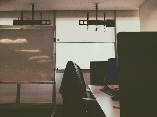 A dimly lit office or classroom space with a desk, computer monitor, keyboard, and a jacket draped over a chair. There is a large whiteboard on the left side, and two projectors mounted on the ceiling. The window blinds are drawn, allowing limited natural light.