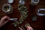A person is rolling a joint on a wooden table. There are cannabis buds scattered across the surface, as well as several glass jars filled with cannabis. A grinder and a few matches are also visible.
