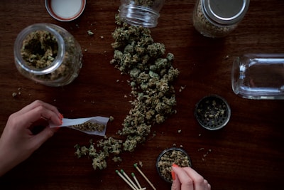 A person is rolling a joint on a wooden table. There are cannabis buds scattered across the surface, as well as several glass jars filled with cannabis. A grinder and a few matches are also visible.