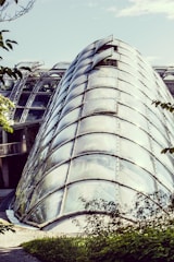 A modern glass structure resembling a large, curved greenhouse. The architecture features transparent panels framed with metal, allowing light to pass through. Surrounding the structure is lush greenery and trees, giving it an organic blend with nature.