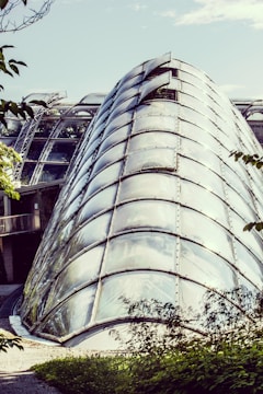 A modern glass structure resembling a large, curved greenhouse. The architecture features transparent panels framed with metal, allowing light to pass through. Surrounding the structure is lush greenery and trees, giving it an organic blend with nature.