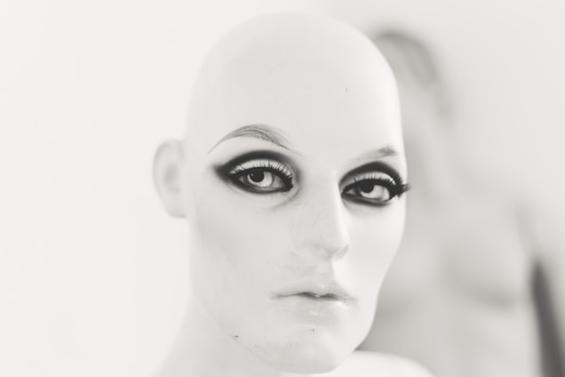 A mannequin head with stylized makeup, featuring dramatic eyeliner and bold eyebrows, set against a blurred background.