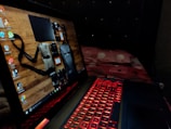 A laptop featuring a red backlit keyboard is situated on a bed with a red and white bedspread. The laptop screen displays a desktop background showing various travel items like a camera and bags against a wooden surface.