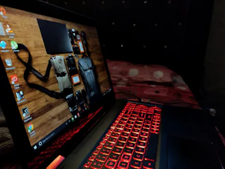A laptop featuring a red backlit keyboard is situated on a bed with a red and white bedspread. The laptop screen displays a desktop background showing various travel items like a camera and bags against a wooden surface.
