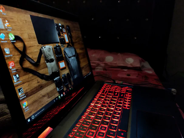 A laptop featuring a red backlit keyboard is situated on a bed with a red and white bedspread. The laptop screen displays a desktop background showing various travel items like a camera and bags against a wooden surface.