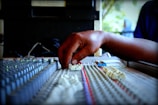 A hand adjusting sliders on an audio mixing console with numerous knobs and faders, indicating a sound engineering environment. The focus is on the hand, suggesting manual control and precision.