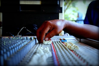 A hand adjusting sliders on an audio mixing console with numerous knobs and faders, indicating a sound engineering environment. The focus is on the hand, suggesting manual control and precision.