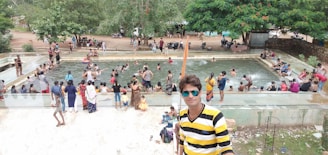 A group of people enjoys a crowded swimming area, with some participants standing and others swimming. The surrounding area has lush green trees and motorbikes parked nearby. A person in a yellow and black striped shirt with sunglasses stands in the foreground holding a stick.