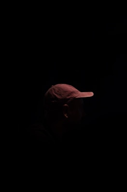 A person wearing a cap is partially illuminated, with the rest of the figure shrouded in darkness, creating a mysterious and dramatic effect.