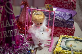 A small doll dressed in a white outfit is seated on a pink swing. The scene is filled with colorful wrapped gifts adorned with intricate patterns. Nearby, a vivid birthday hat with tinsel and 'Happy Birthday' text is visible. The table is scattered with confetti, and there's also a decorated picture frame containing a photograph.