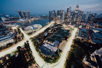 The image features a cityscape at dusk, showcasing a densely packed skyline with tall skyscrapers lit up against the evening sky. In the foreground, brightly illuminated roads curve through the city, surrounded by lush greenery and various structures. A large body of water is visible, along with several iconic modern architectural structures.