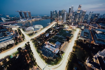 The image features a cityscape at dusk, showcasing a densely packed skyline with tall skyscrapers lit up against the evening sky. In the foreground, brightly illuminated roads curve through the city, surrounded by lush greenery and various structures. A large body of water is visible, along with several iconic modern architectural structures.