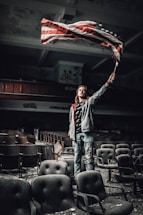 A person stands among rows of worn, dusty theater seats in an abandoned or dilapidated auditorium. The individual holds and waves an American flag with a determined expression, surrounded by graffiti-covered walls and peeling paint.