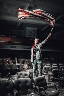 A person stands among rows of worn, dusty theater seats in an abandoned or dilapidated auditorium. The individual holds and waves an American flag with a determined expression, surrounded by graffiti-covered walls and peeling paint.