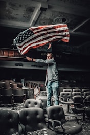 A person is holding a large American flag inside an abandoned auditorium with worn-out and dusty seats. The ceiling shows signs of decay and the atmosphere is dimly lit. The person is wearing a jacket and jeans, standing among rows of empty chairs.