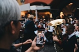 A person is taking a photo of a brass band performance using a smartphone. The band members are wearing uniforms and playing musical instruments such as trumpets and tubas. The scene is crowded, possibly at an outdoor event, with people gathered closely together.