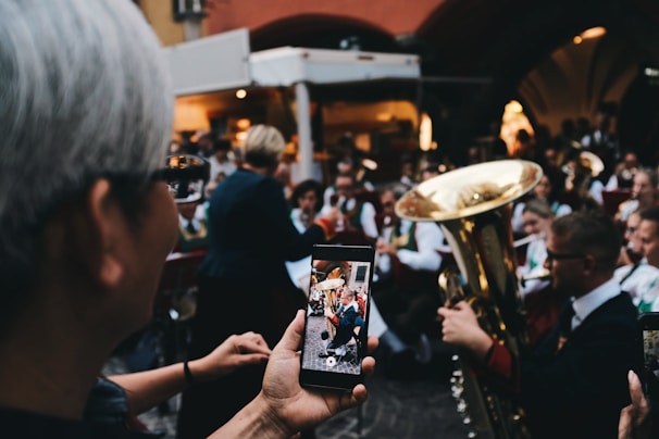 A person is taking a photo of a brass band performance using a smartphone. The band members are wearing uniforms and playing musical instruments such as trumpets and tubas. The scene is crowded, possibly at an outdoor event, with people gathered closely together.