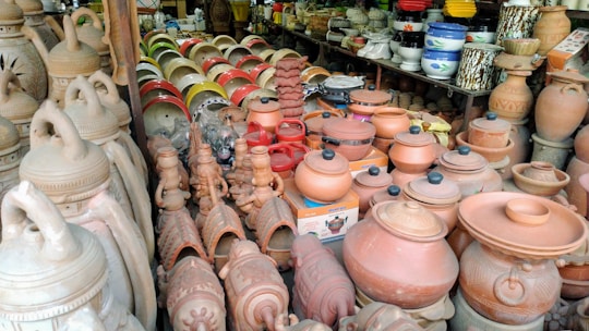 A variety of clay pots and kitchenware are displayed on shelves and tables, featuring different shapes and sizes. Terracotta pottery is prominently featured, with some pieces decorated with intricate patterns. The setting appears to be a market or shop, with colorful plastic bowls visible in the background.