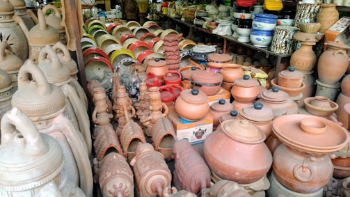 A variety of clay pots and kitchenware are displayed on shelves and tables, featuring different shapes and sizes. Terracotta pottery is prominently featured, with some pieces decorated with intricate patterns. The setting appears to be a market or shop, with colorful plastic bowls visible in the background.