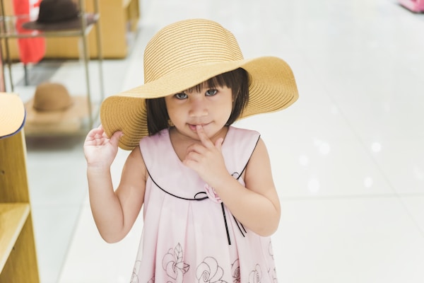 A young child wearing a wide-brimmed straw hat and a light pink dress with a floral pattern stands in a brightly lit store. The child is looking at the camera with a thoughtful expression.