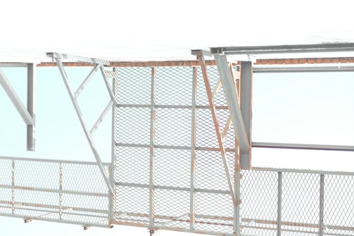 A metallic structure with a mesh-style railing and horizontal and diagonal beams. The background features a bright and overexposed sky, suggesting an industrial or construction setting.