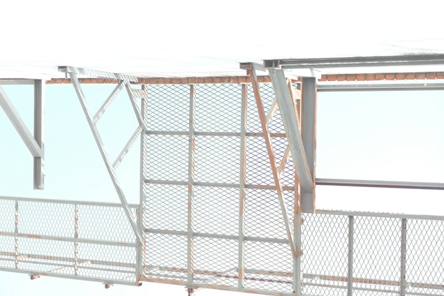 A metallic structure with a mesh-style railing and horizontal and diagonal beams. The background features a bright and overexposed sky, suggesting an industrial or construction setting.