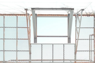 A metal structure with a pattern of crisscrossed diamond shapes creates a grid-like appearance. The open frame allows light to pass through, casting shadows and reflections. The metal appears to have a slight rust on the edges, giving an industrial feel.