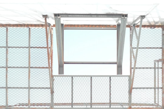 A metal structure with a pattern of crisscrossed diamond shapes creates a grid-like appearance. The open frame allows light to pass through, casting shadows and reflections. The metal appears to have a slight rust on the edges, giving an industrial feel.