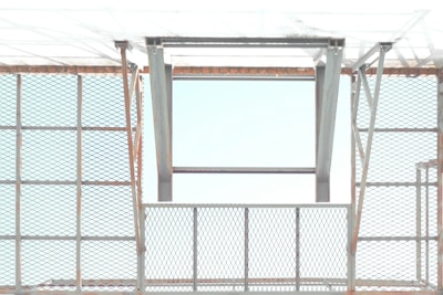 A metal structure with a pattern of crisscrossed diamond shapes creates a grid-like appearance. The open frame allows light to pass through, casting shadows and reflections. The metal appears to have a slight rust on the edges, giving an industrial feel.
