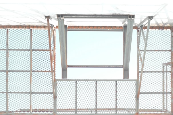 A metal structure with a pattern of crisscrossed diamond shapes creates a grid-like appearance. The open frame allows light to pass through, casting shadows and reflections. The metal appears to have a slight rust on the edges, giving an industrial feel.