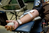 A person is lying down with their arm resting on a padded surface, donating blood. A blood pressure cuff is wrapped around the upper arm, and a needle is inserted with tape securing it in place. The hand holds a stress ball to aid blood flow.
