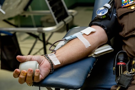 A person is lying down with their arm resting on a padded surface, donating blood. A blood pressure cuff is wrapped around the upper arm, and a needle is inserted with tape securing it in place. The hand holds a stress ball to aid blood flow.