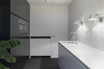 A modern kitchen with sleek, minimalist design features. A white countertop and backsplash paired with dark cabinets create a stark contrast. Wall-mounted lights provide illumination, and a built-in oven is present in the left-hand cabinetry. A green plant adds a touch of nature.
