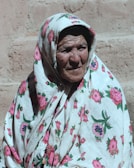 An elderly person wearing a floral-patterned headscarf and robe is captured in front of a textured wall with gentle sunlight casting shadows.
