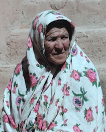 An elderly person wearing a floral-patterned headscarf and robe is captured in front of a textured wall with gentle sunlight casting shadows.