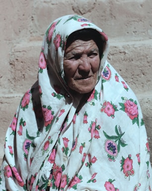 An elderly person wearing a floral-patterned headscarf and robe is captured in front of a textured wall with gentle sunlight casting shadows.