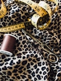 Detailed view of a sewing setup including a leopard print fabric, a yellow measuring tape, a spool of brown thread, and a pair of metal scissors.