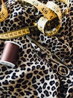 Detailed view of a sewing setup including a leopard print fabric, a yellow measuring tape, a spool of brown thread, and a pair of metal scissors.