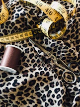 Detailed view of a sewing setup including a leopard print fabric, a yellow measuring tape, a spool of brown thread, and a pair of metal scissors.